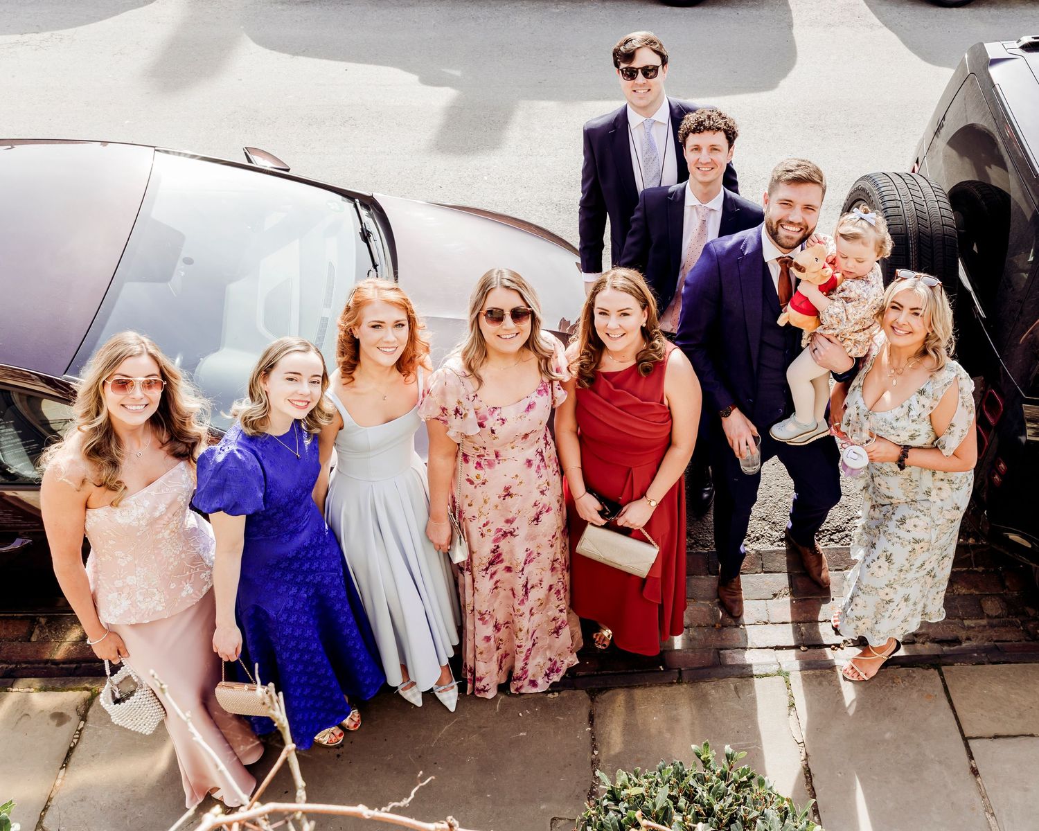 Group of wedding guests posing together in formal attire beside a black vehicle.