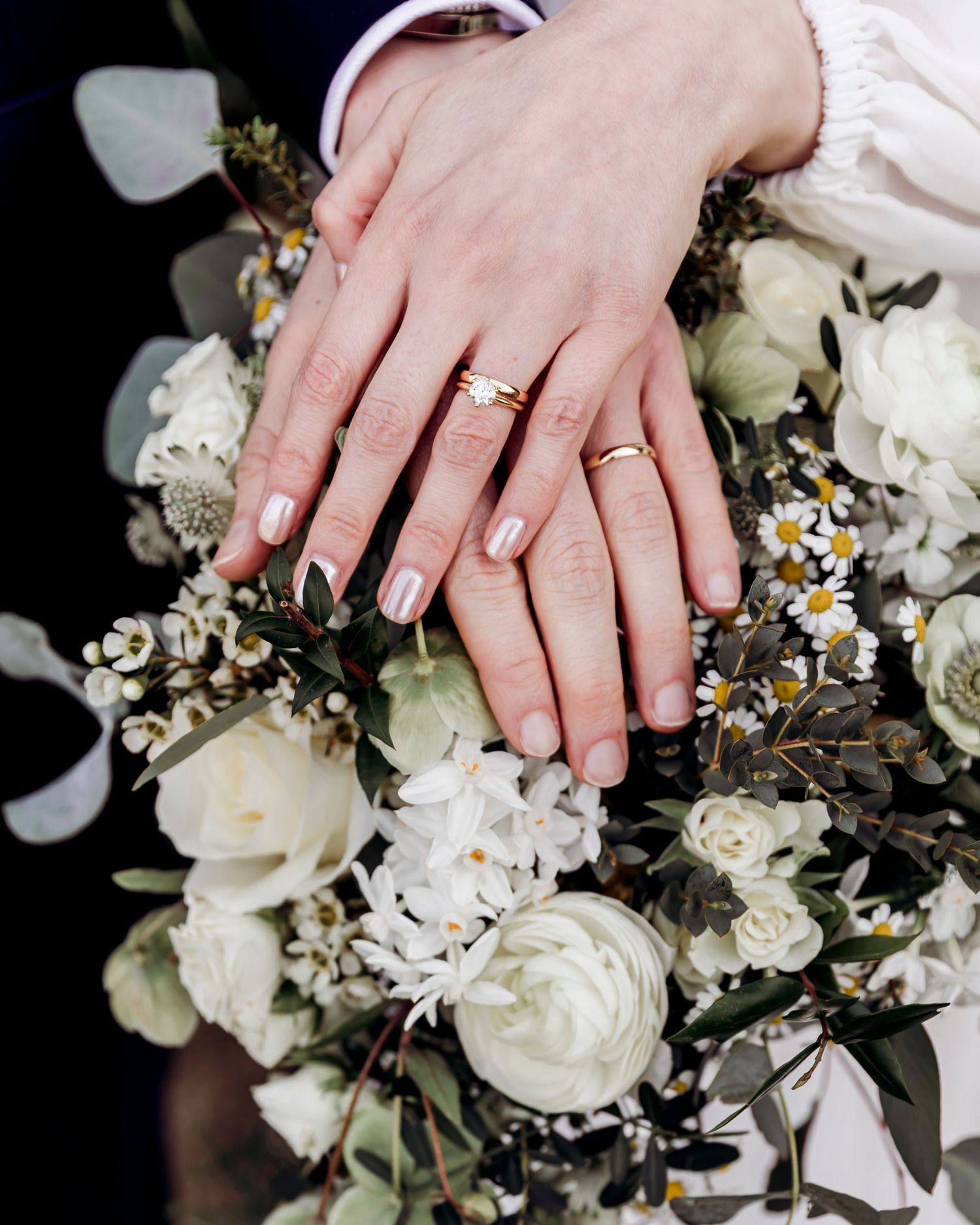 Newlyweds display gold wedding rings on hands resting over white floral bouquet.