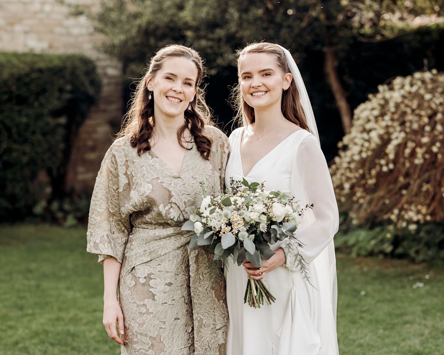 Bride in white gown holding bouquet poses with bridesmaid in gold dress outdoors.