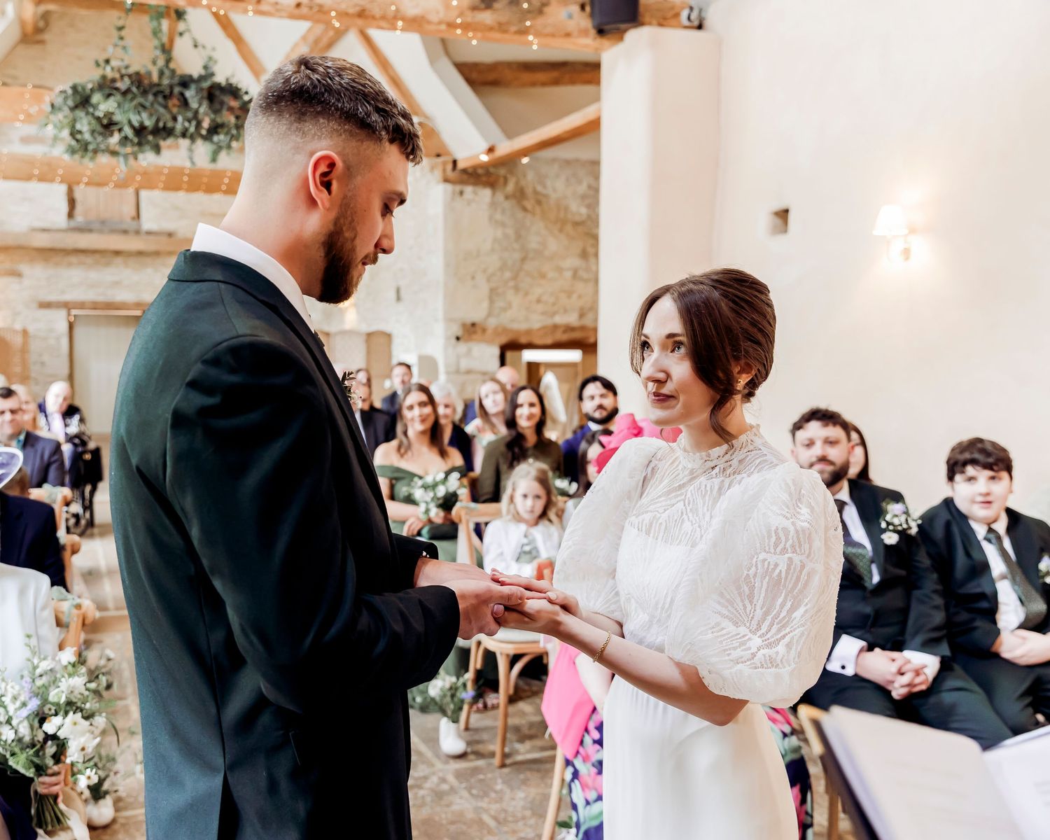 Bride and groom exchange rings during an intimate wedding ceremony in a rustic barn venue with guests seated behind.