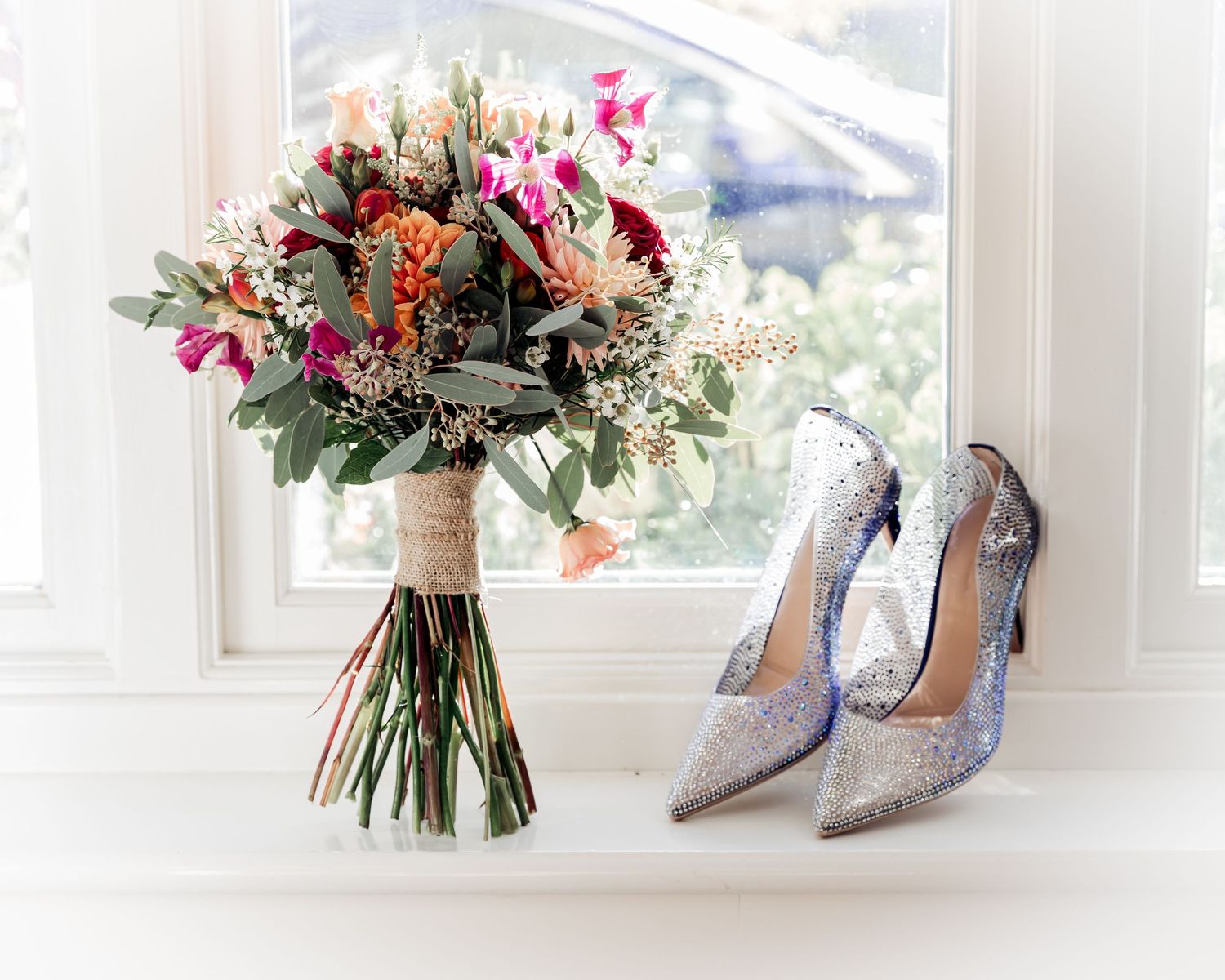 Bridal bouquet of colorful wildflowers beside sparkling silver stiletto heels on a white windowsill.