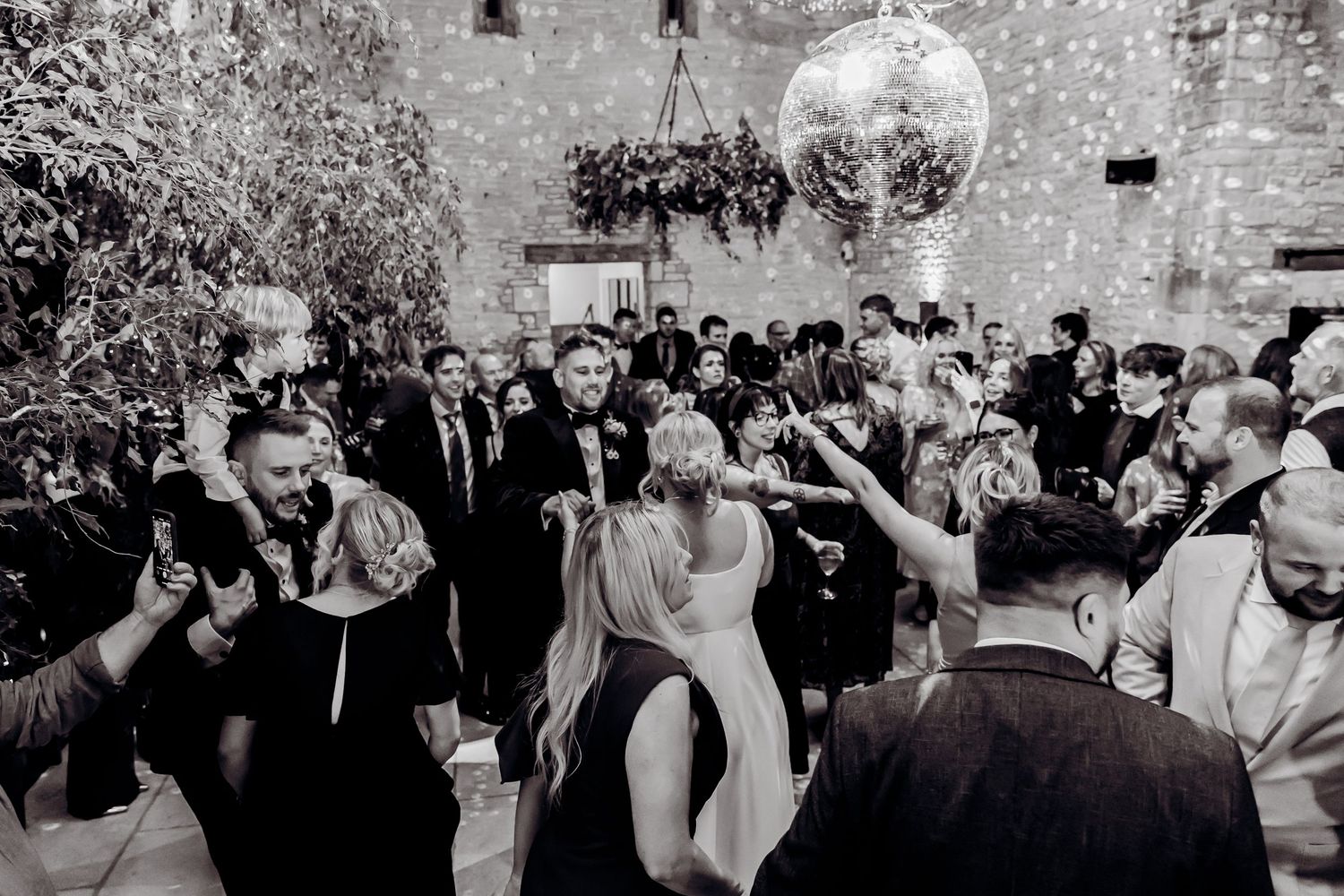 Black and white photo of wedding guests dancing under disco ball at lively reception party.