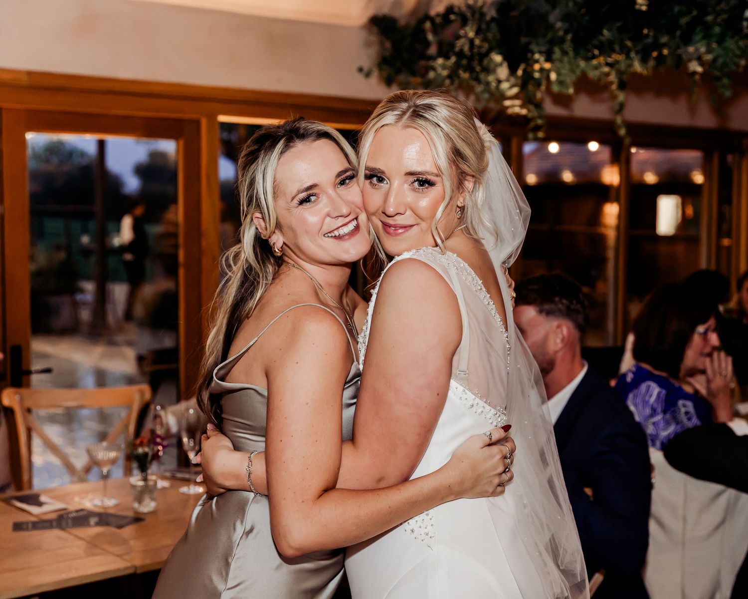 Bride and bridesmaid embrace warmly at a rustic indoor wedding reception.