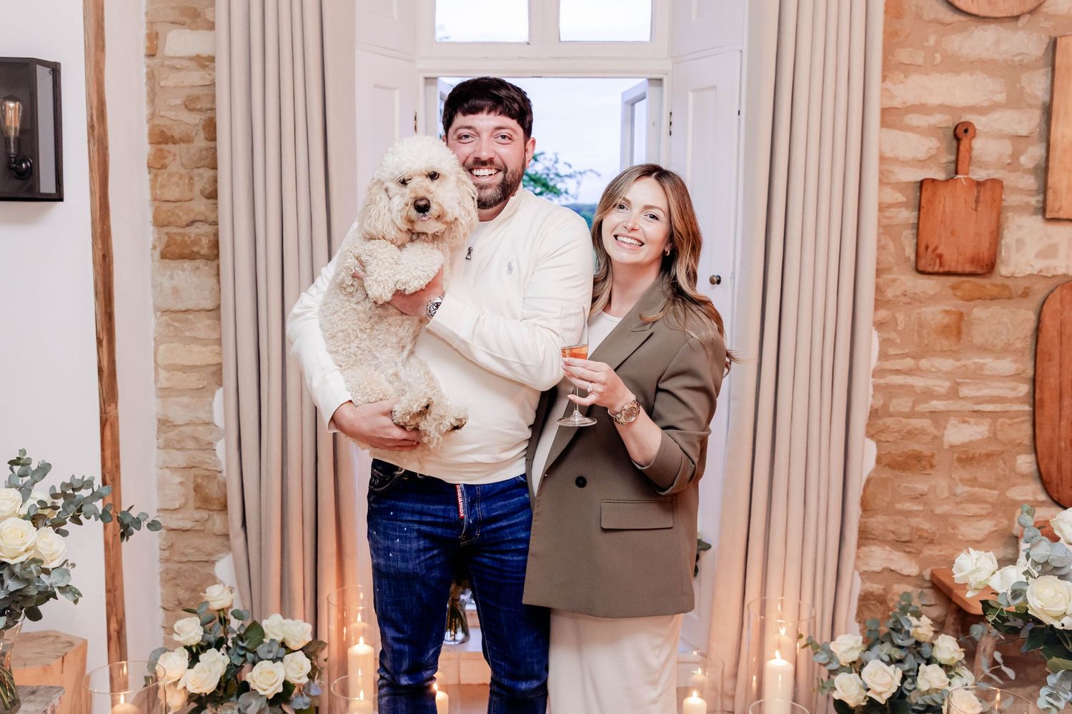 A smiling couple holding a fluffy white dog and champagne glass, standing in a decorated doorway with flowers.