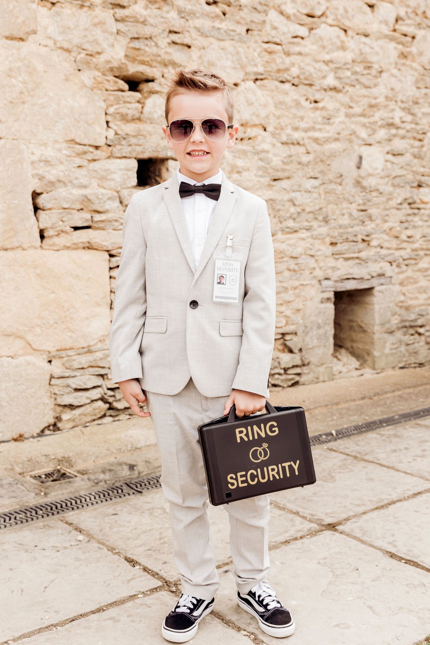 Young ring bearer in gray suit, bow tie, and sunglasses holding a black Ring Security briefcase at a wedding.