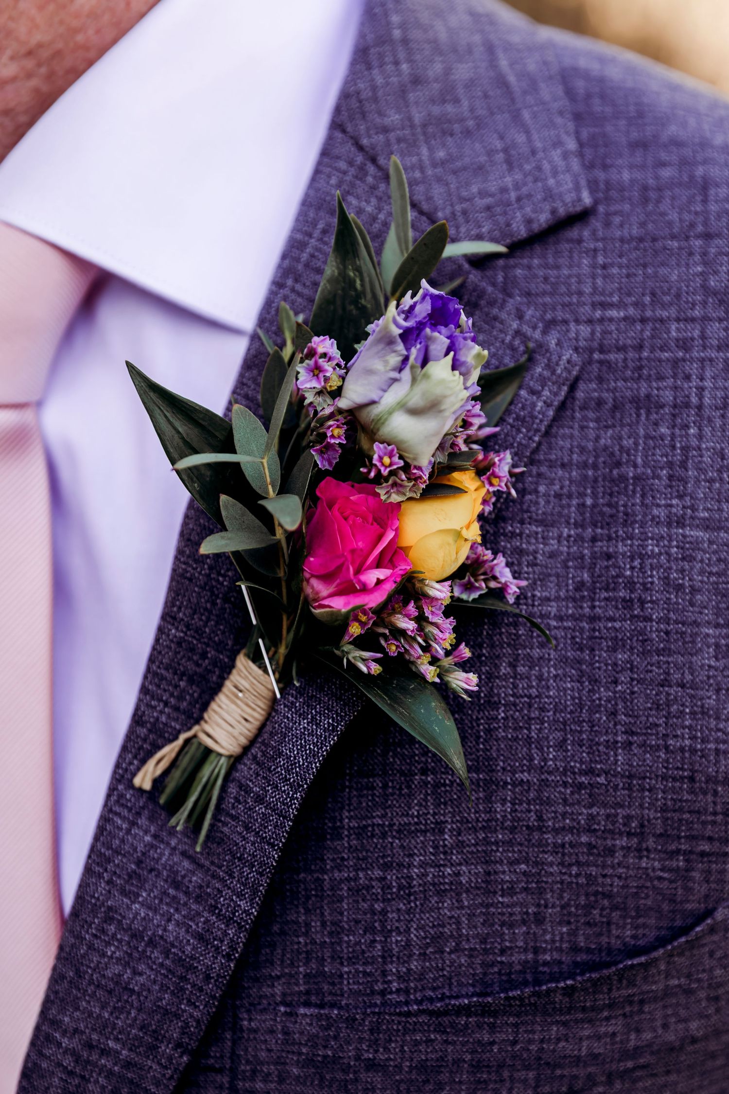 Colorful wedding boutonniere with pink, yellow, and purple flowers on a grey textured suit lapel.