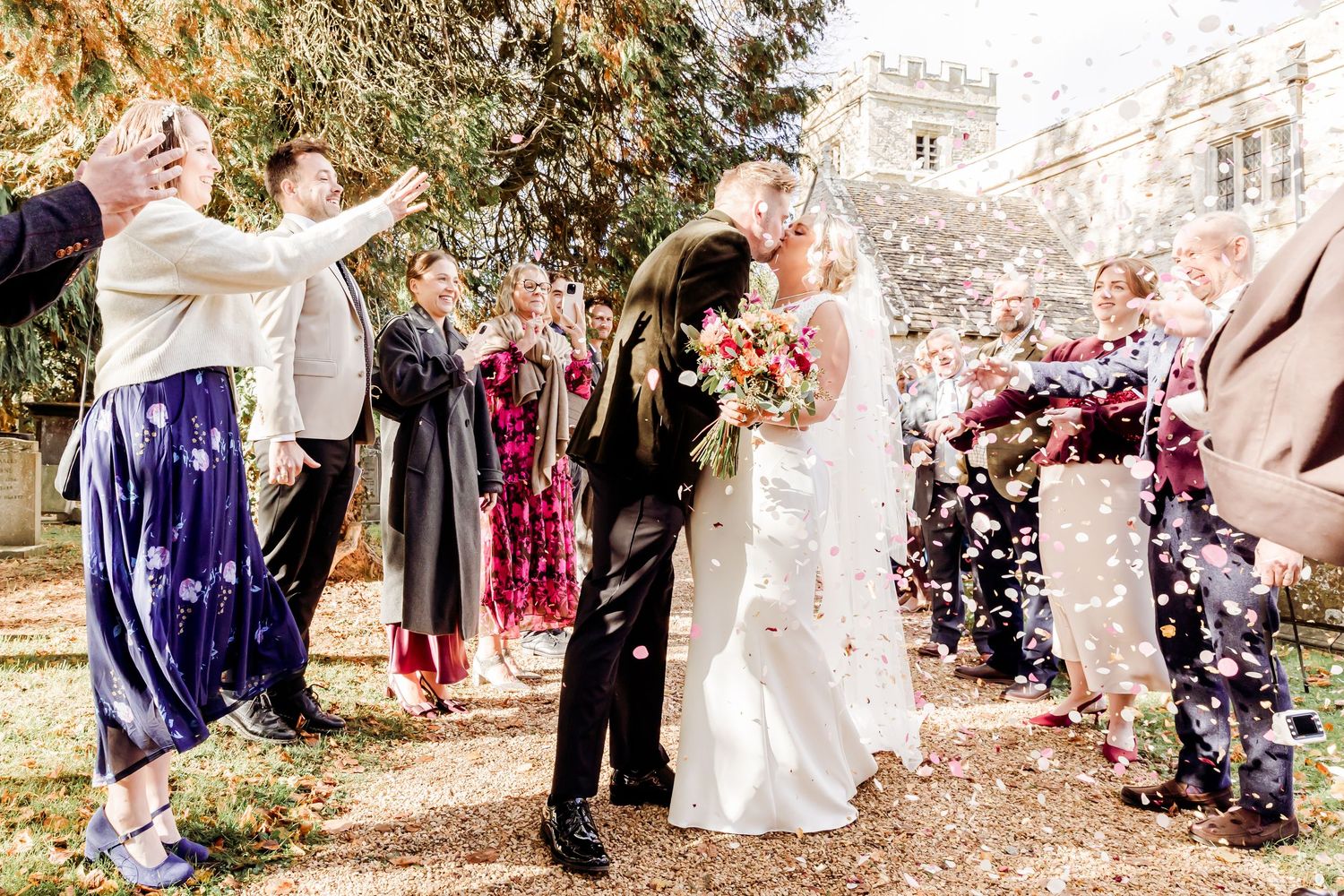Newlyweds kiss outdoors as guests toss colorful confetti while someone captures the moment on phone.