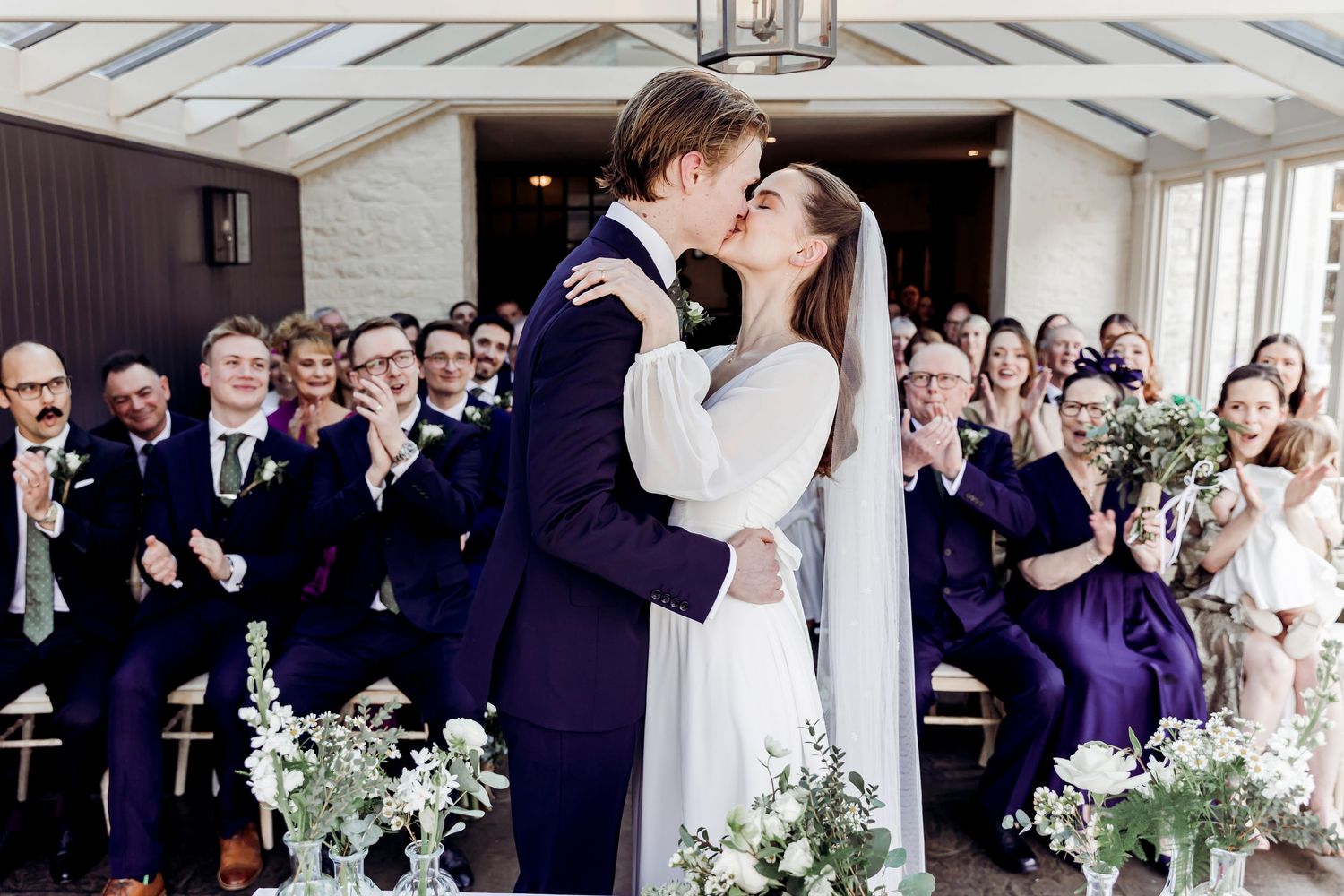 Bride and groom share first kiss at wedding altar surrounded by cheering guests.