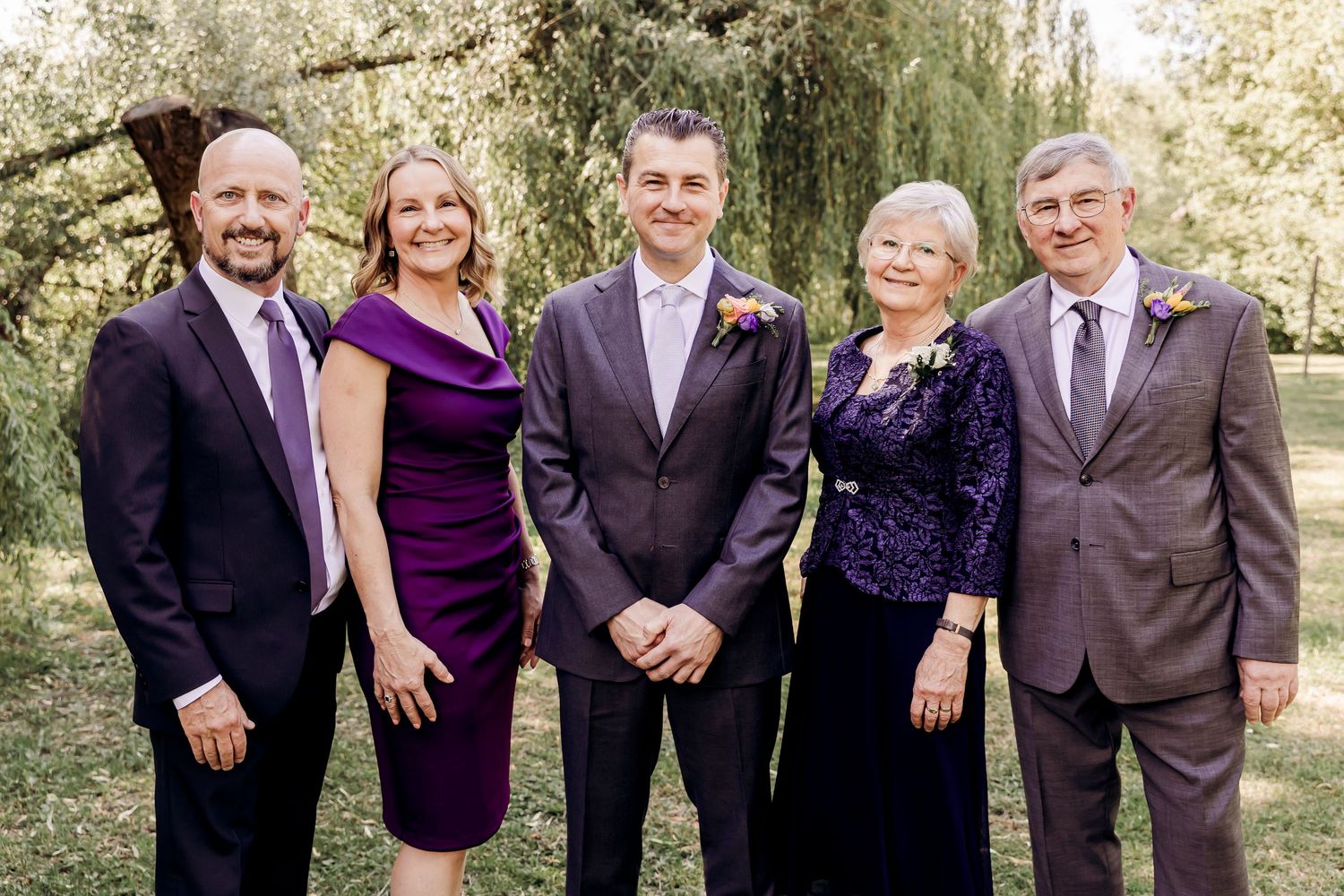 Family group portrait at outdoor wedding with five adults in formal purple and gray attire among green trees.
