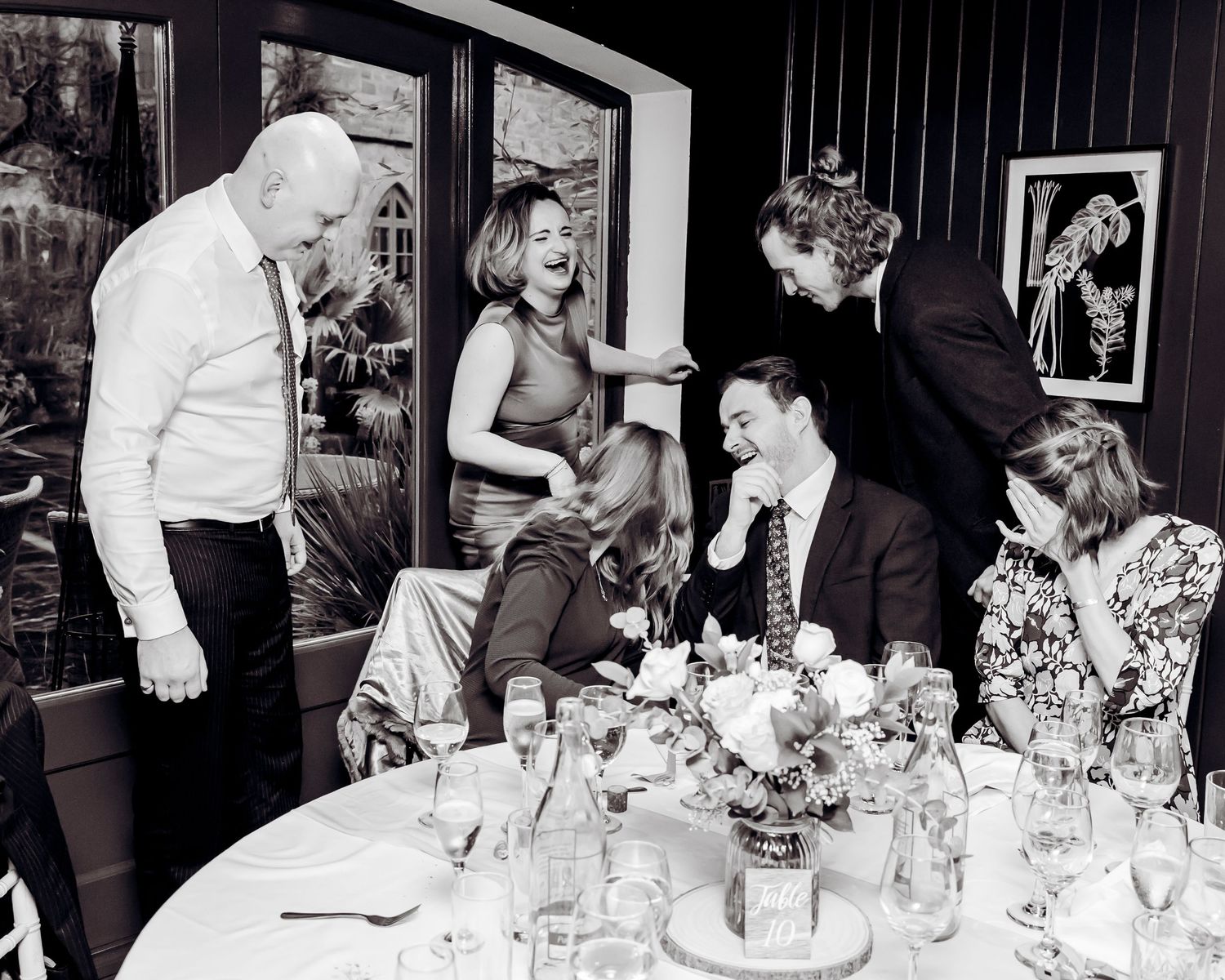 Black and white photo of joyful wedding guests laughing together around a reception table.