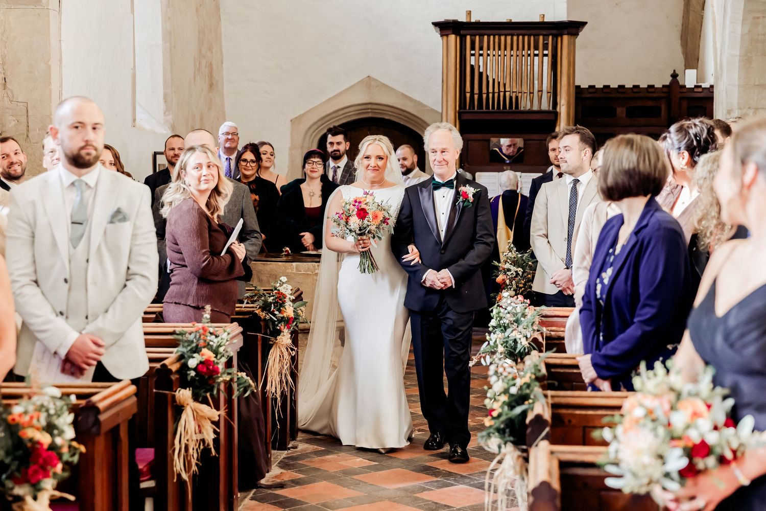 Bride walking down church aisle with father past seated wedding guests during ceremony procession.