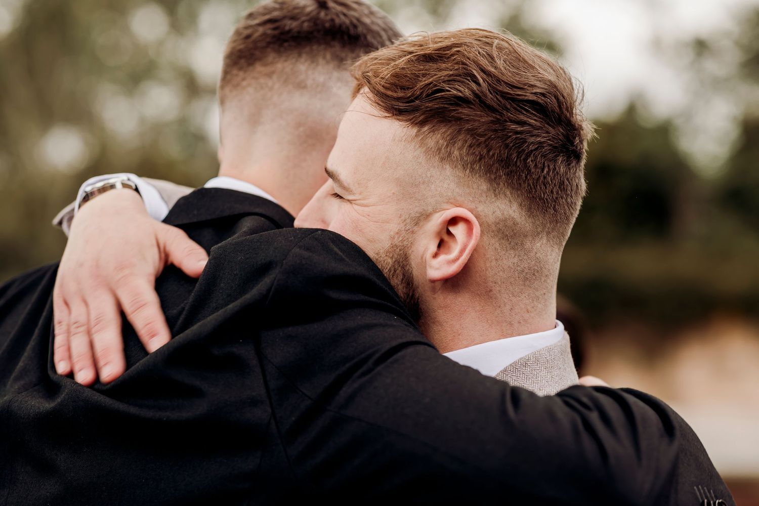 Two young men in formal suits share a warm embrace outdoors, captured in a tender moment.
