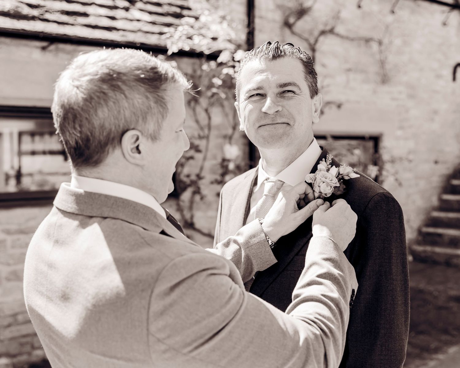 Black and white photo of two grooms adjusting boutonnieres at an outdoor wedding ceremony.