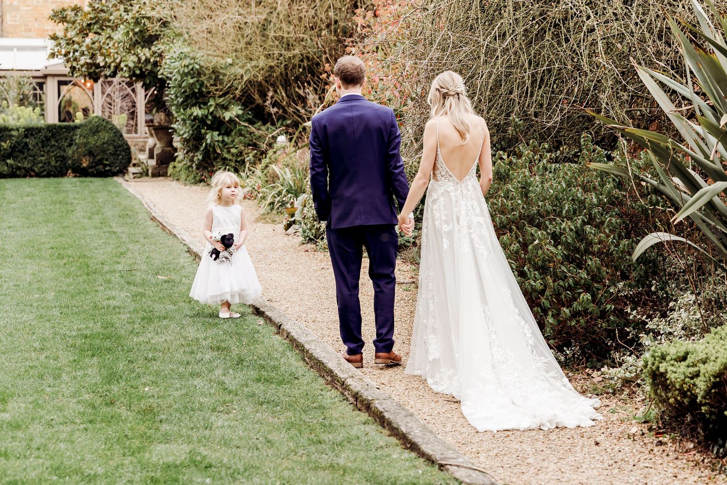 Bride, groom, and flower girl walking along a garden path at an elegant wedding venue.