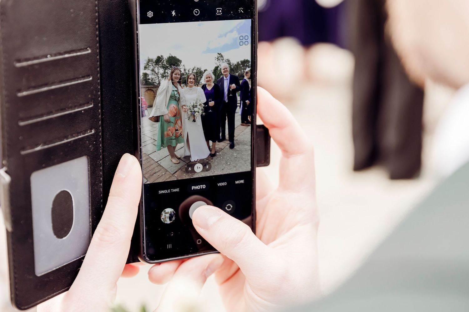Person photographing a group of wedding guests on a smartphone with a black leather wallet case at an outdoor event.