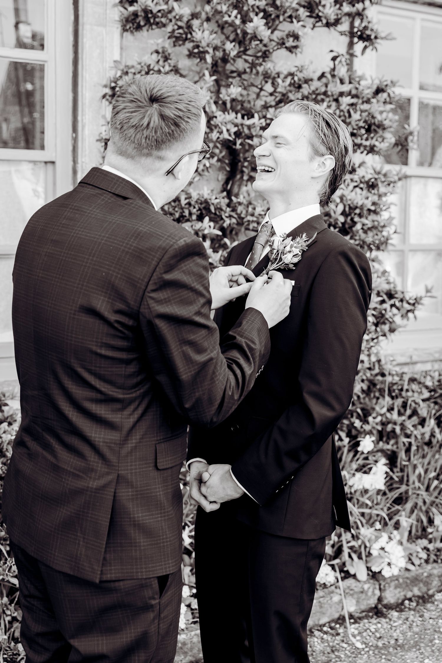 Best man pinning a boutonniere on a laughing groom in a dark suit outdoors in black and white.