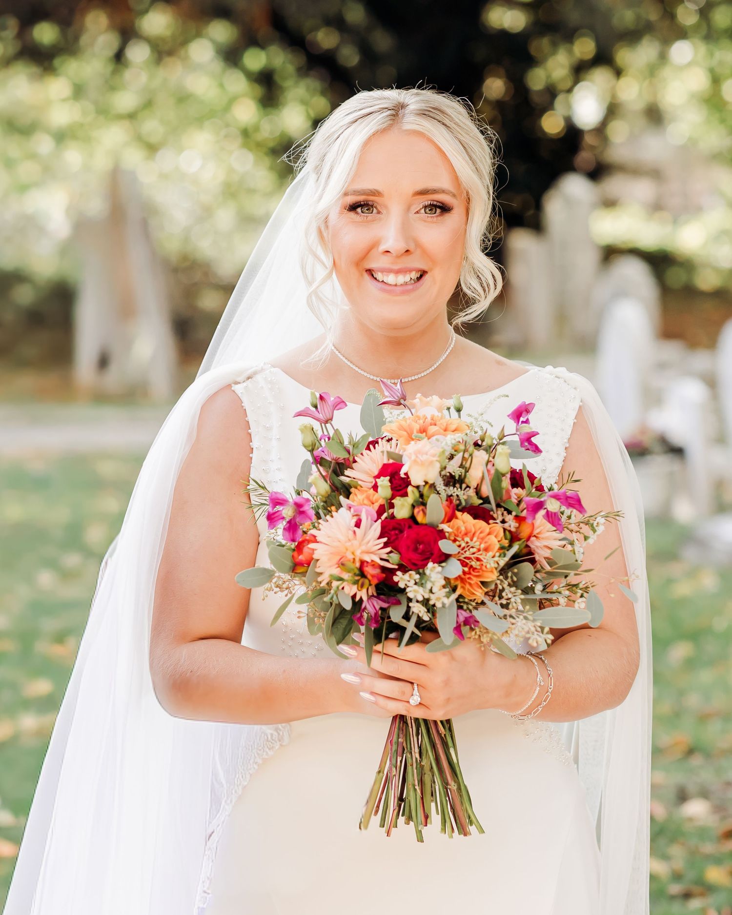 Smiling bride in white gown and veil holding a vibrant bouquet of orange, pink, and red flowers outdoors.