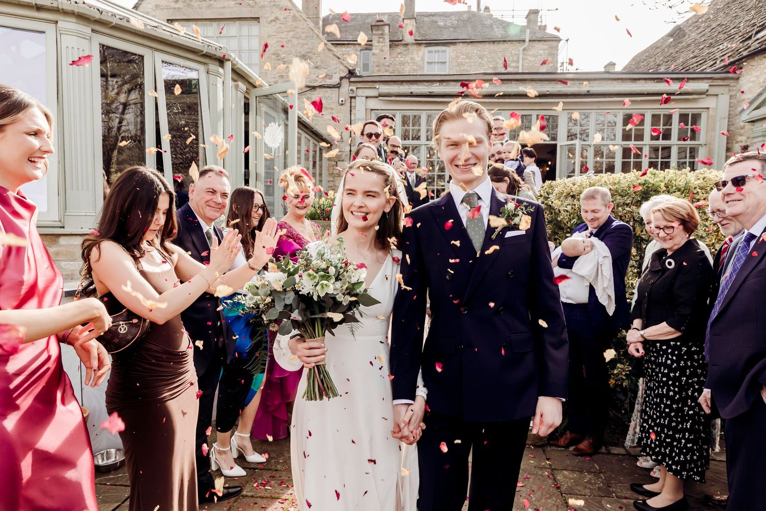 Bride and groom walk through confetti shower outside stone wedding venue with guests.
