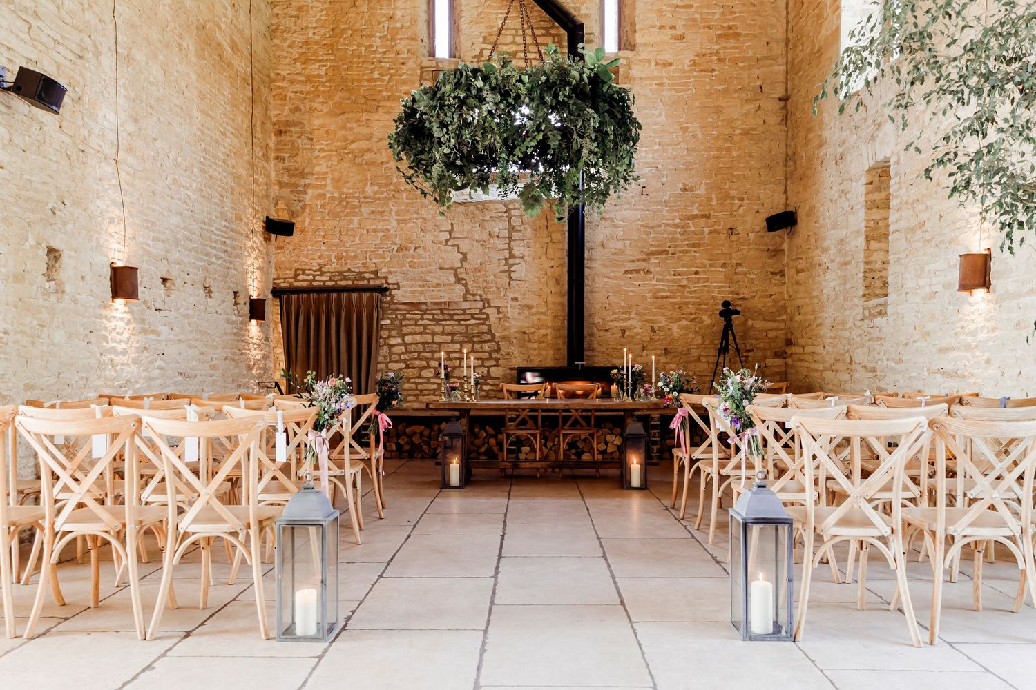 Rustic barn wedding ceremony venue with wooden cross-back chairs, lanterns, and hanging floral installation.
