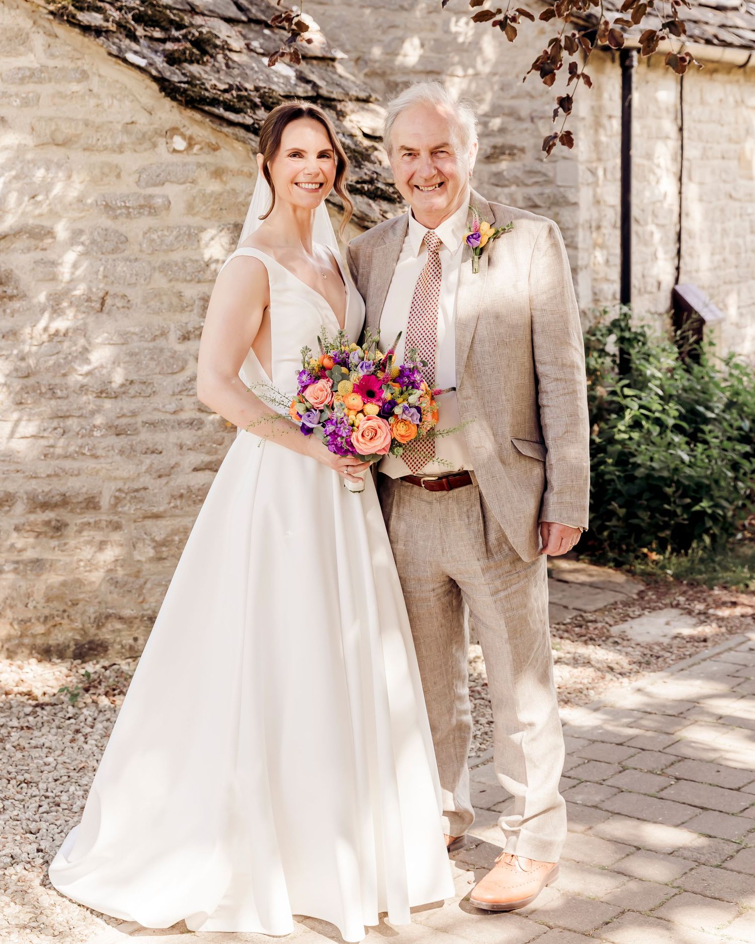 Bride in white gown holds colorful bouquet beside older man in tan suit outside stone building.