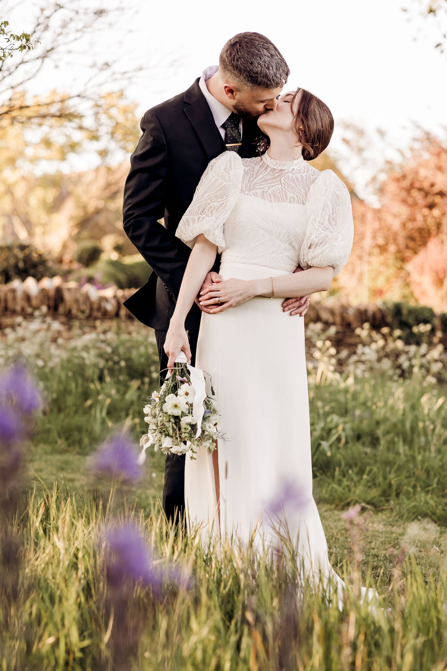 Couple shares a romantic kiss in a field of purple wildflowers, bride holding a delicate white bouquet.