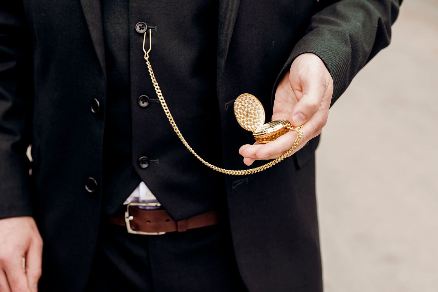 Man in black suit holding open gold pocket watch with chain attached to lapel pin.