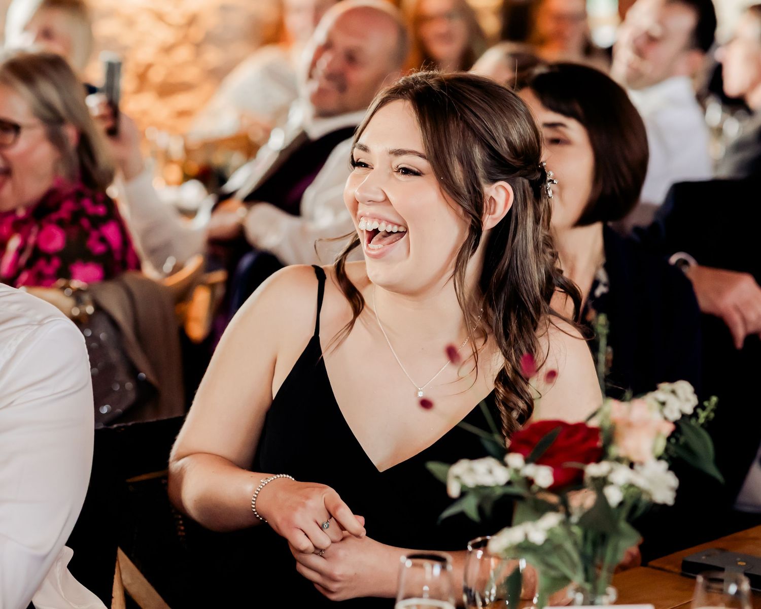 A woman in a black dress laughs joyfully at a wedding reception dinner table.