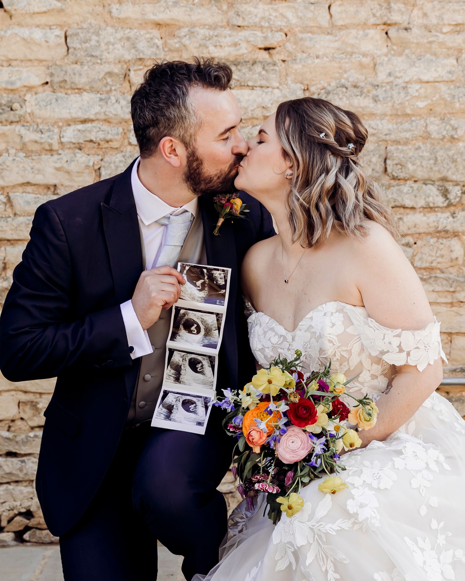 Couple kisses while groom holds ultrasound photos, bride carries vibrant bouquet at wedding.