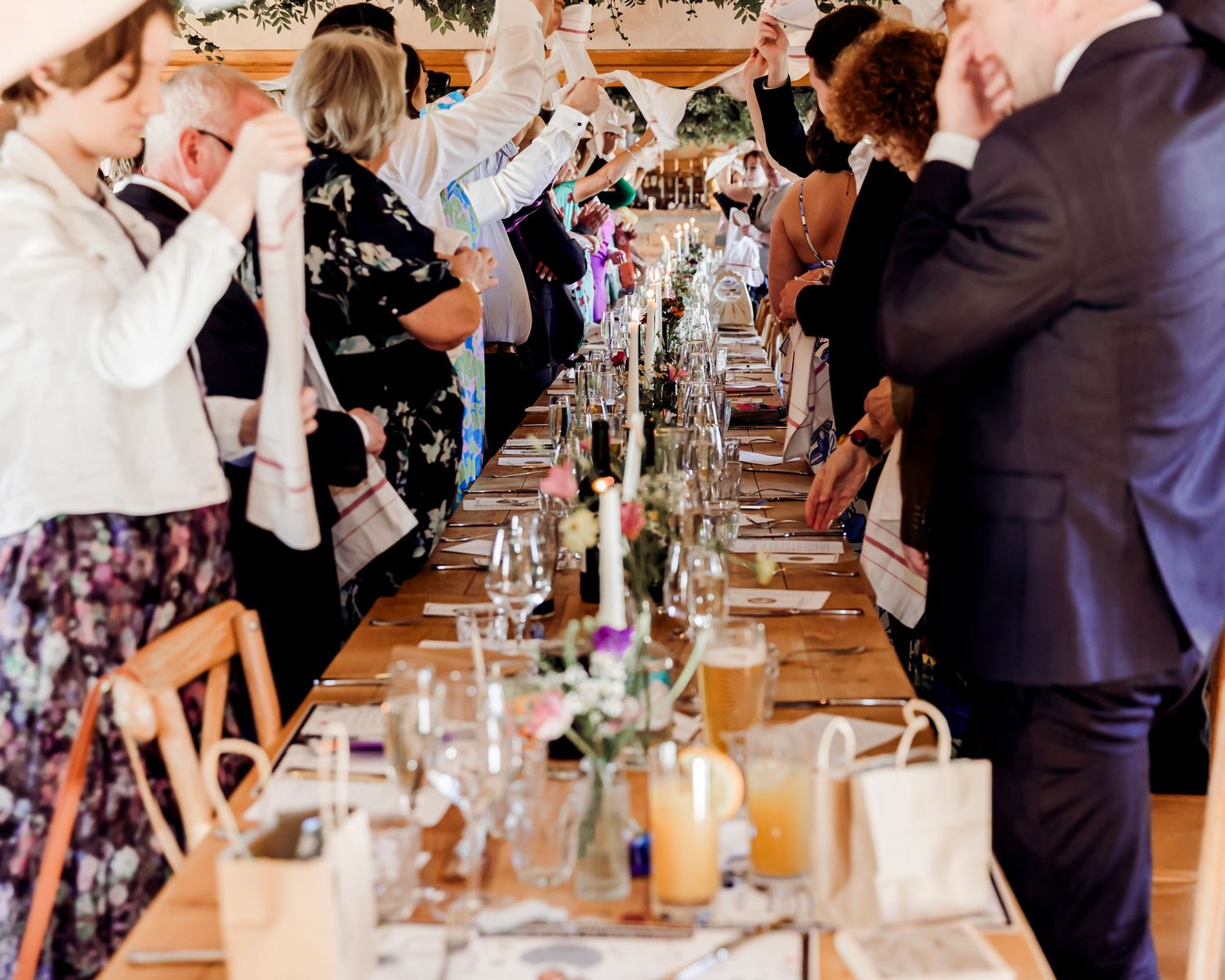 Wedding guests raise phones and cheer as bride and groom walk between long decorated reception tables.