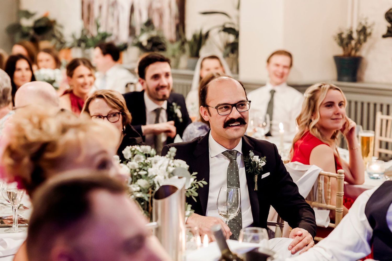 Wedding guest wearing novelty mustache prop laughing during reception speeches.