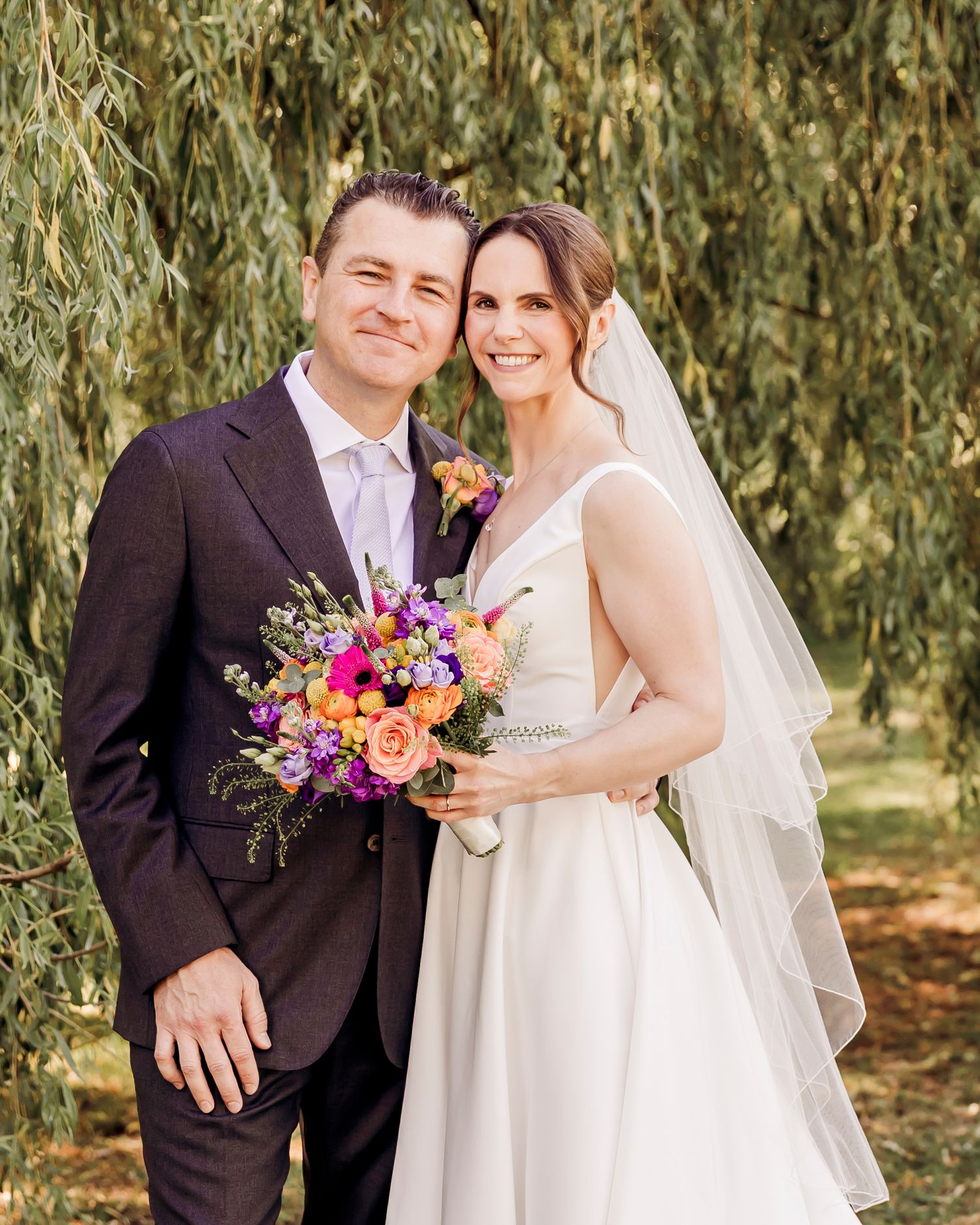 A smiling bride and groom pose together outdoors under a willow tree, bride holding a colorful bouquet of orange and purple flowers.