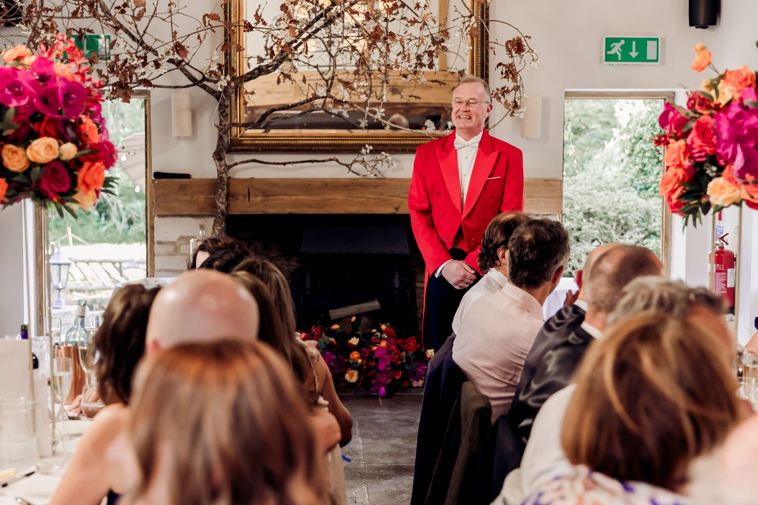 Man in red jacket addresses wedding guests seated before fireplace adorned with colorful floral arrangements.