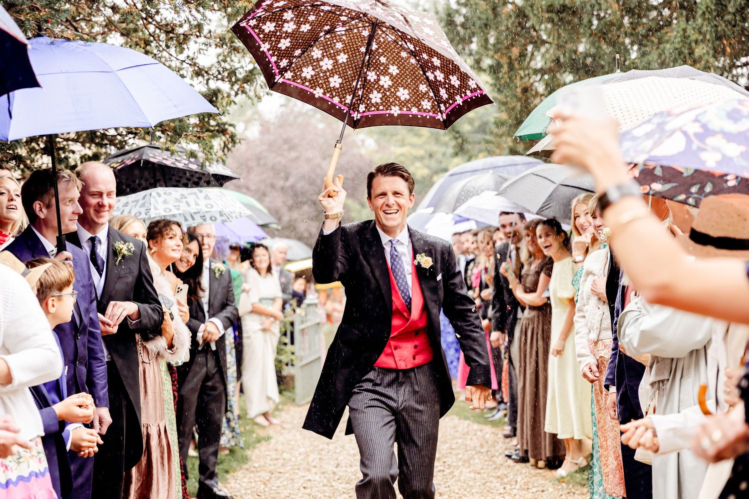 Groom walks joyfully down outdoor wedding aisle holding floral umbrella in the rain, guests lining both sides.