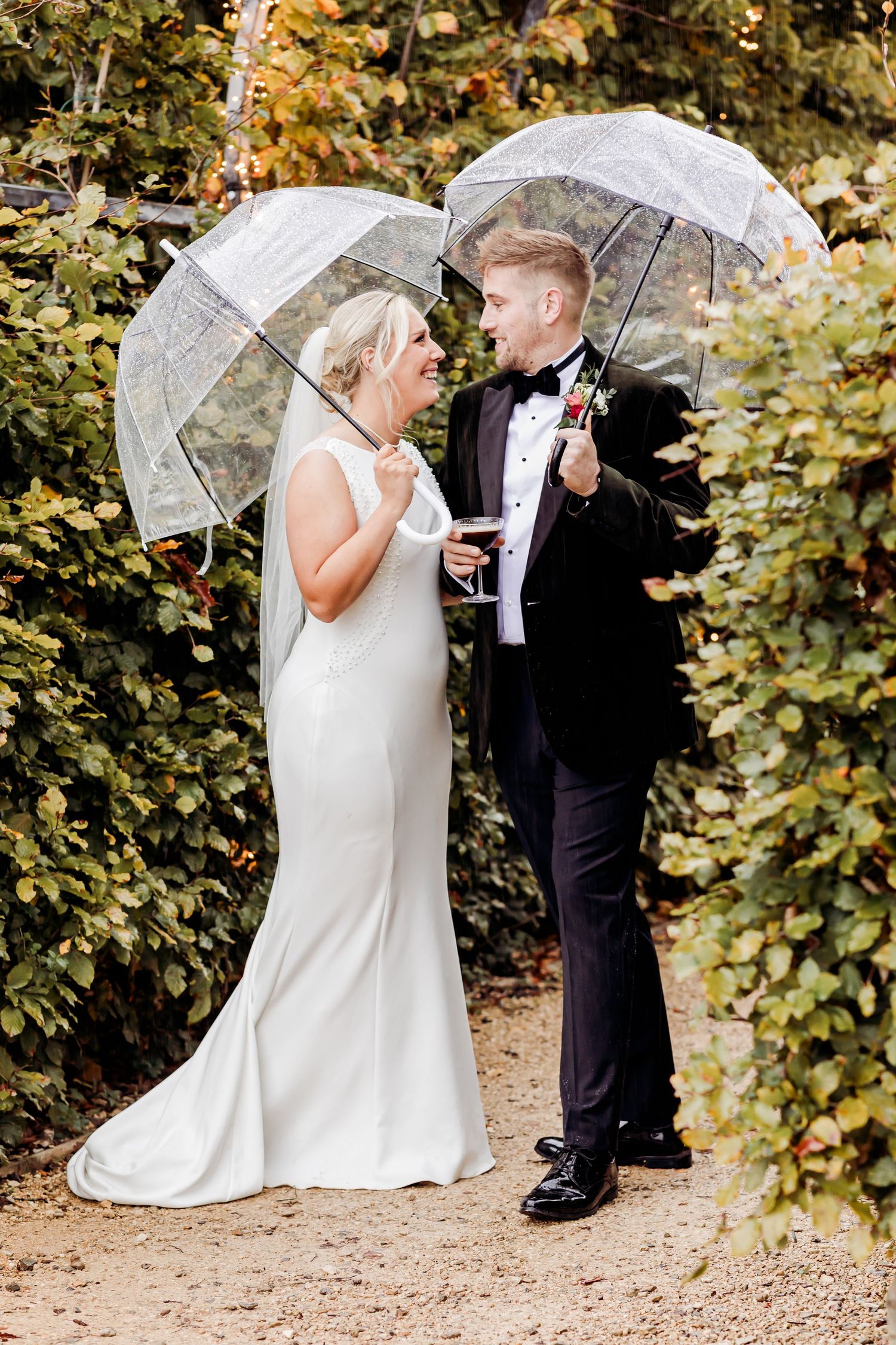 Bride and groom smile at each other under clear umbrellas surrounded by autumn foliage.