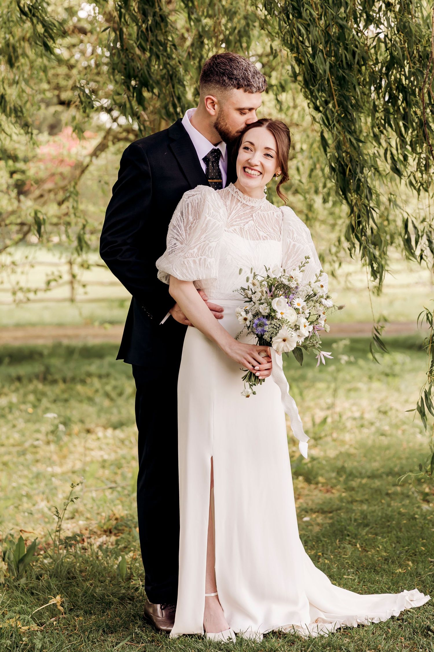 Groom in black suit kissing bride in white lace dress holding wildflower bouquet outdoors.
