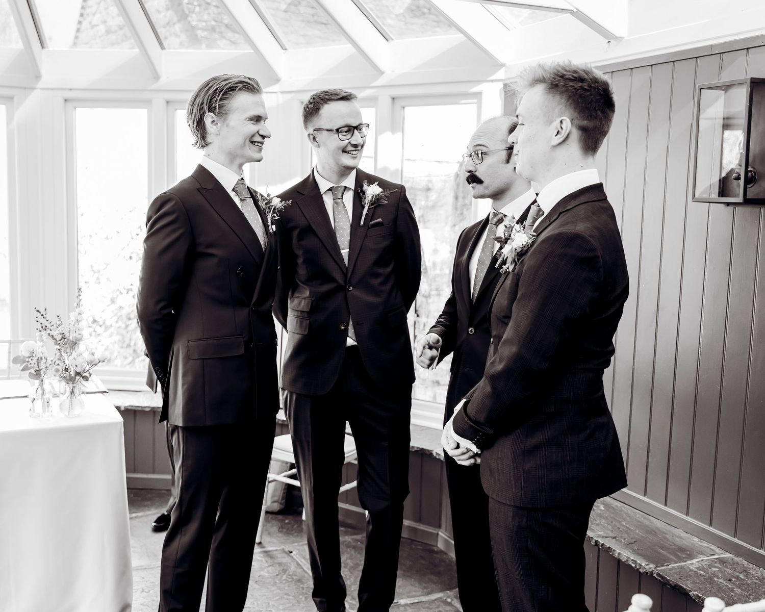 Group of groomsmen in dark suits laughing together in a bright conservatory in black and white.