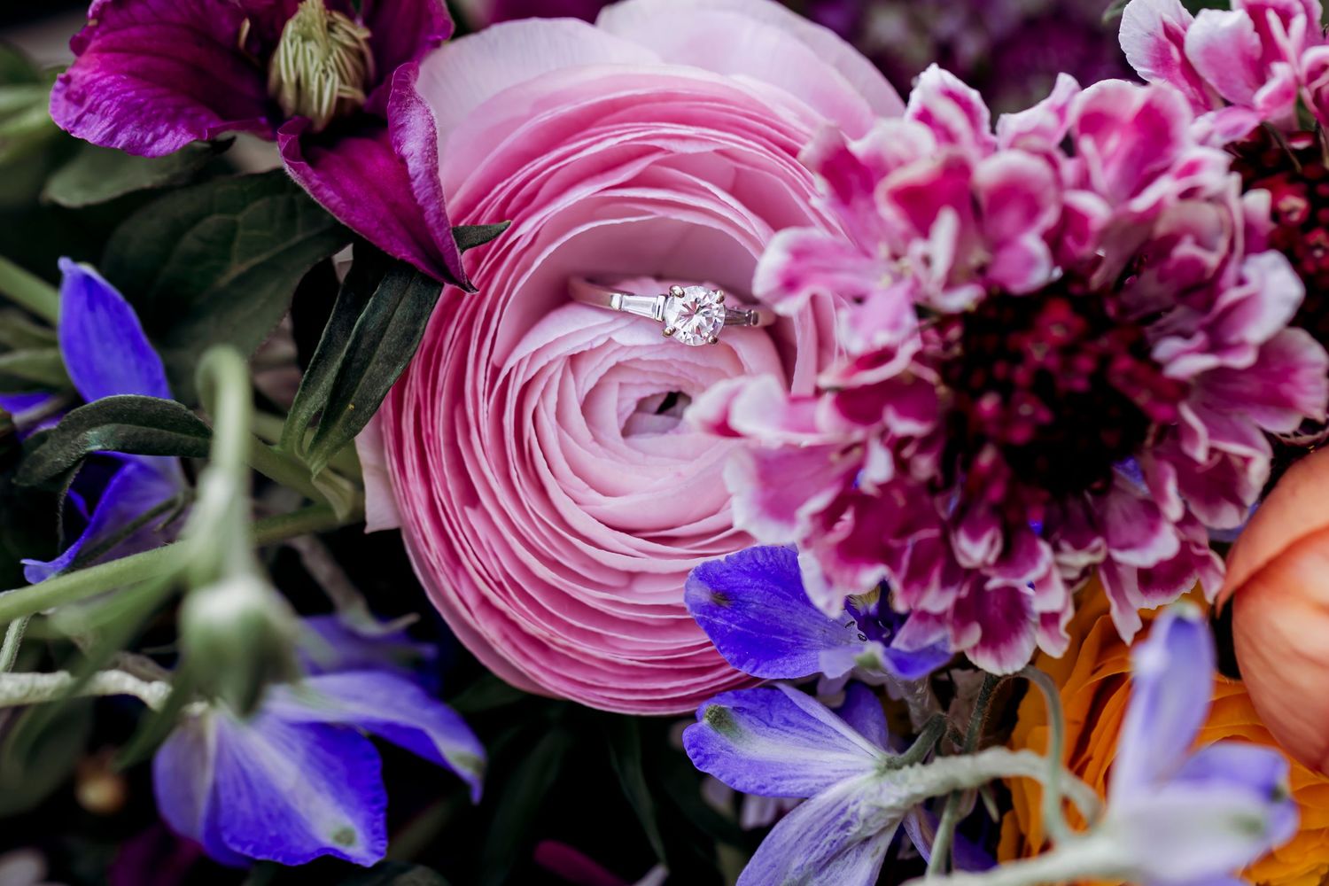 Diamond engagement ring nestled in vibrant pink and purple wedding bouquet flowers.