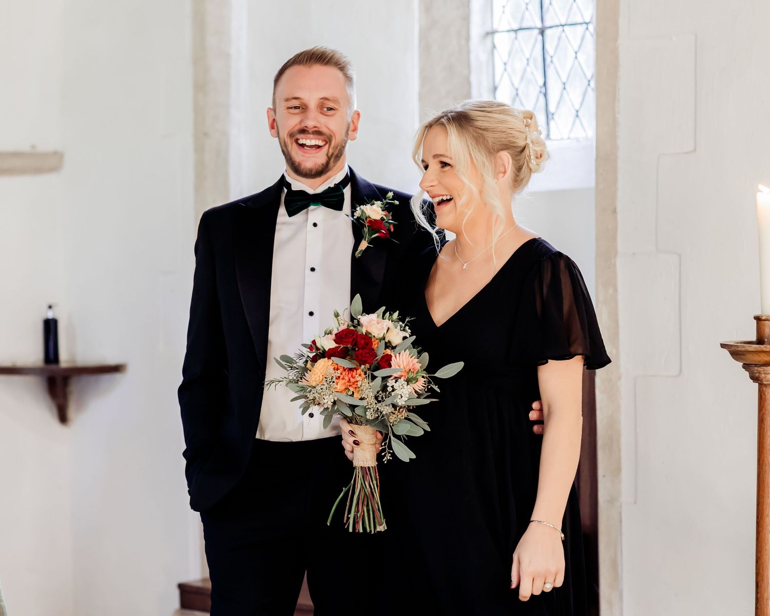 Smiling man in black tuxedo and woman in black dress holding a colorful bridal bouquet in a church.