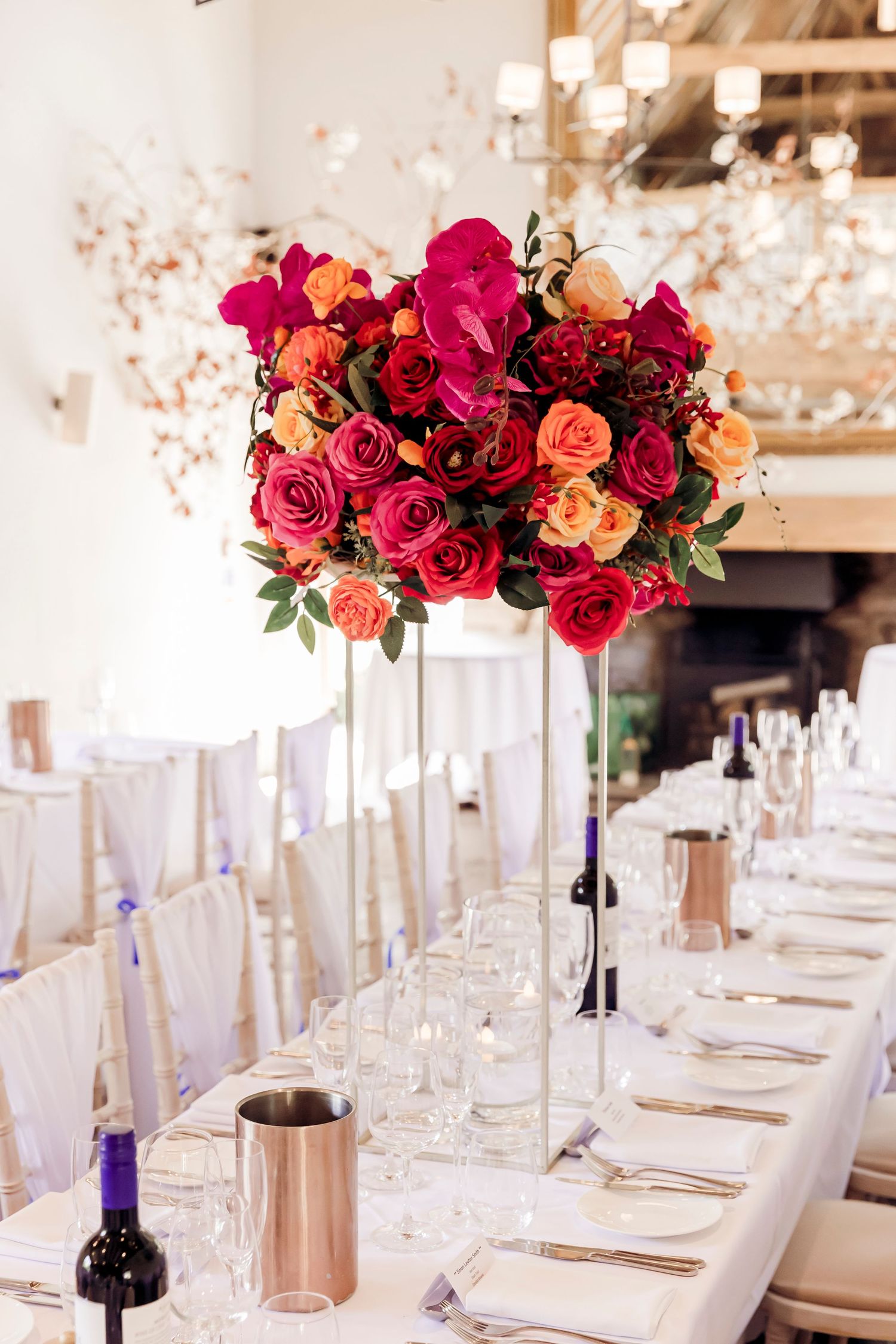 Tall glass vase centerpiece with vibrant red, pink, and orange roses on a white wedding reception table.