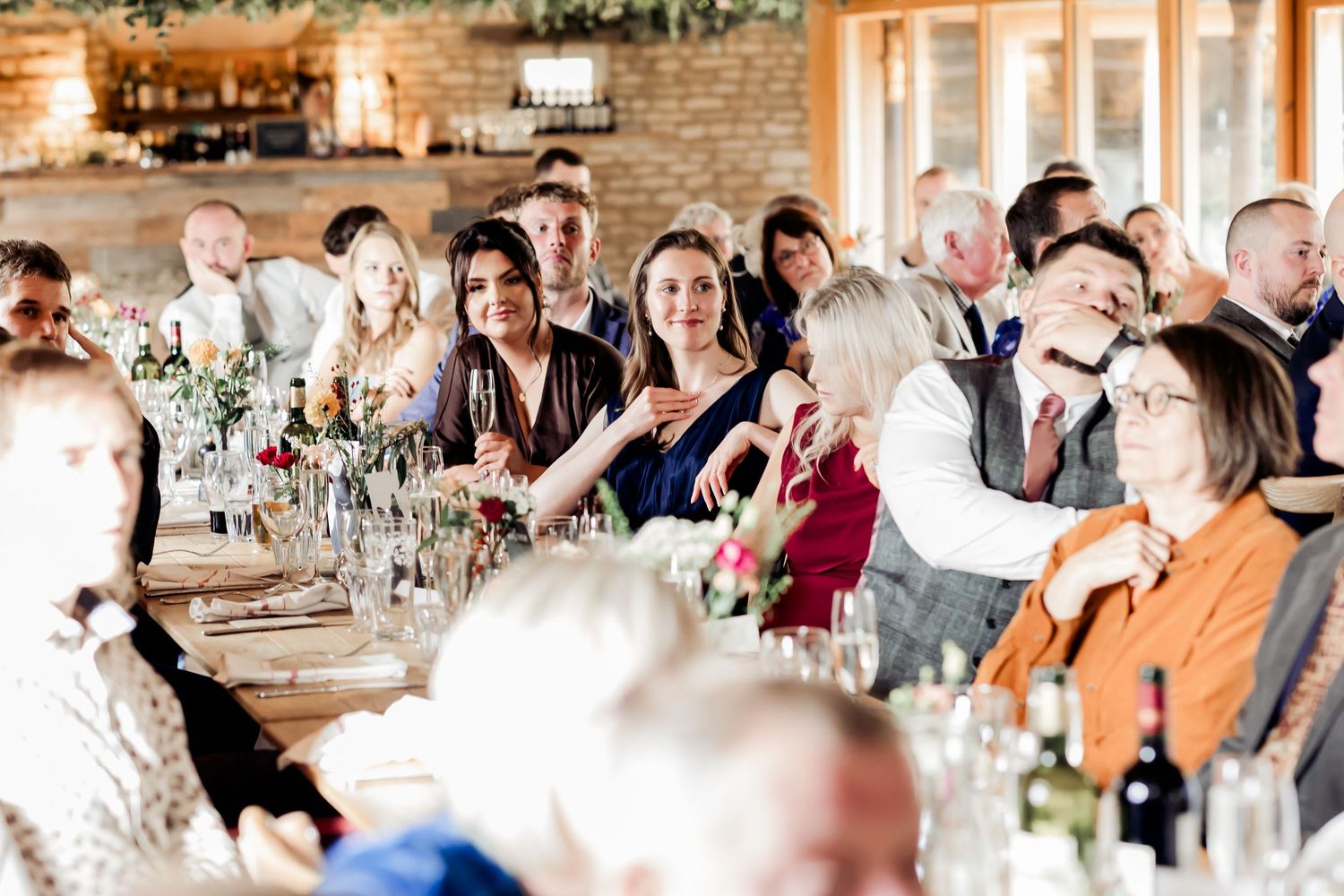 Wedding guests smile and mingle at a long decorated reception table in a rustic venue.
