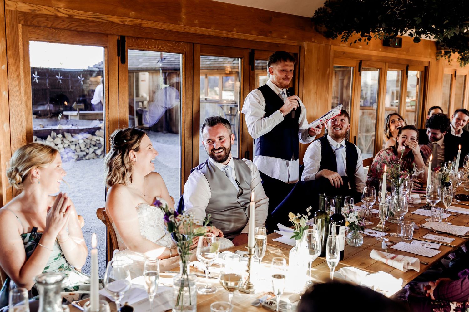 Wedding guests laughing during a heartfelt speech at an elegant rustic reception dinner.