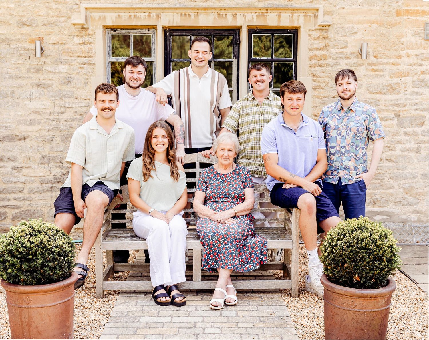 Family group portrait of eight people posing on stone steps outside a traditional stone building with potted topiary.