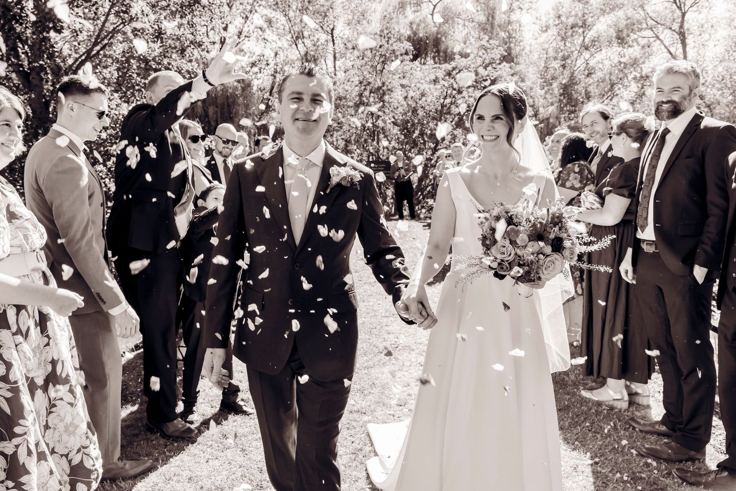 Newlyweds walk through confetti shower in black and white photo as guests celebrate outdoor wedding ceremony.