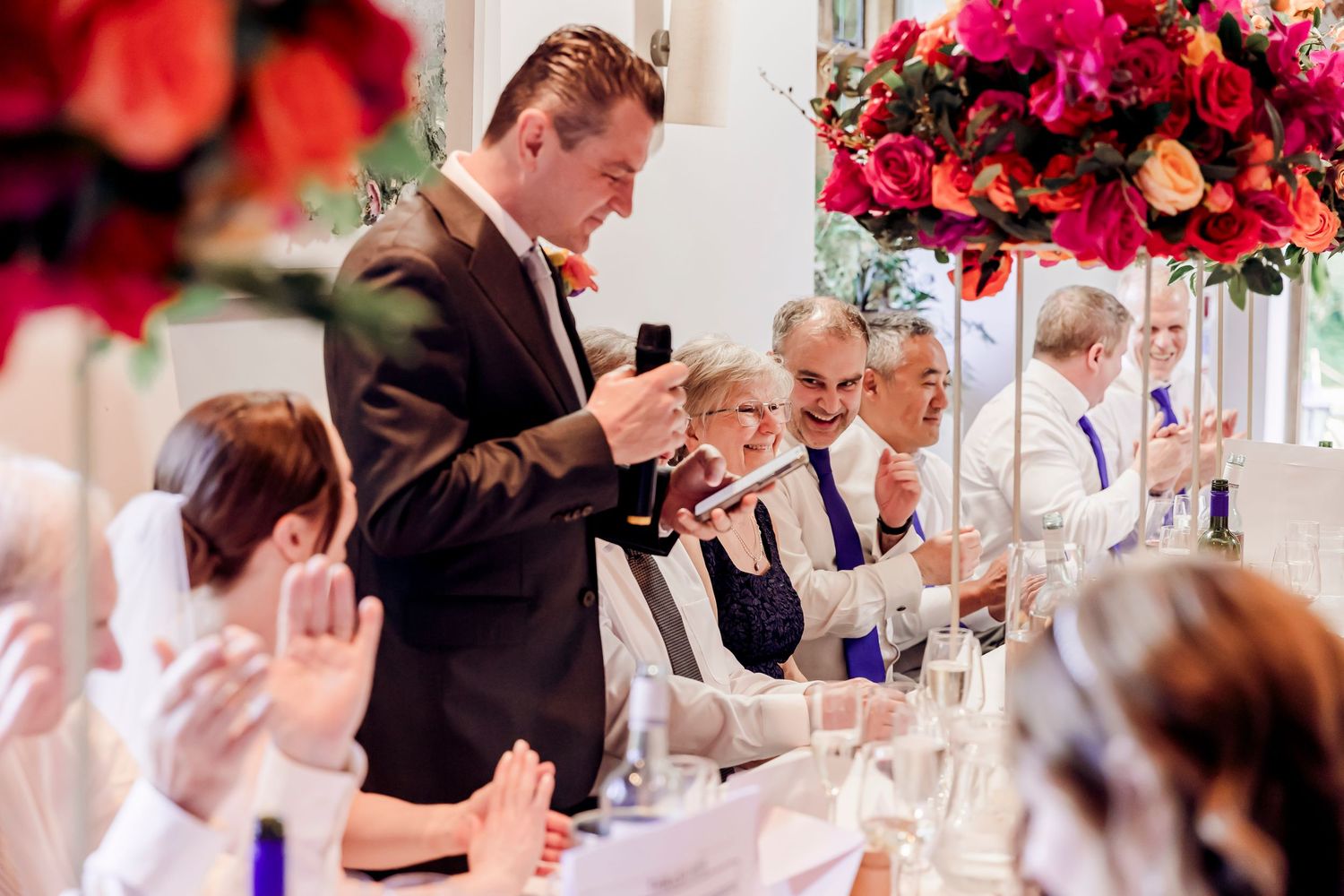 A groom delivering a speech at a wedding reception surrounded by guests and floral centerpieces.