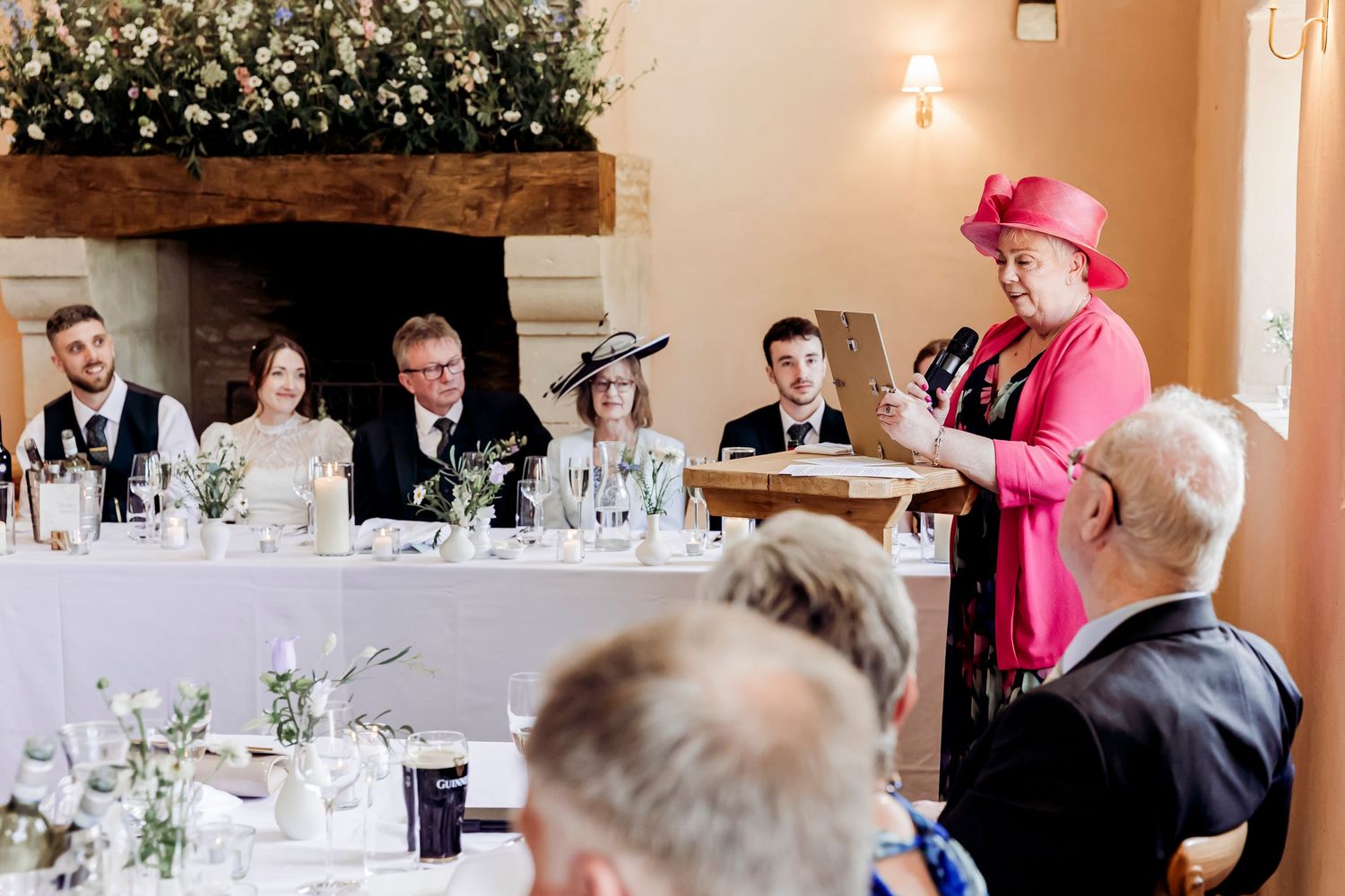 Woman in pink hat and suit giving a speech at an elegant wedding reception.