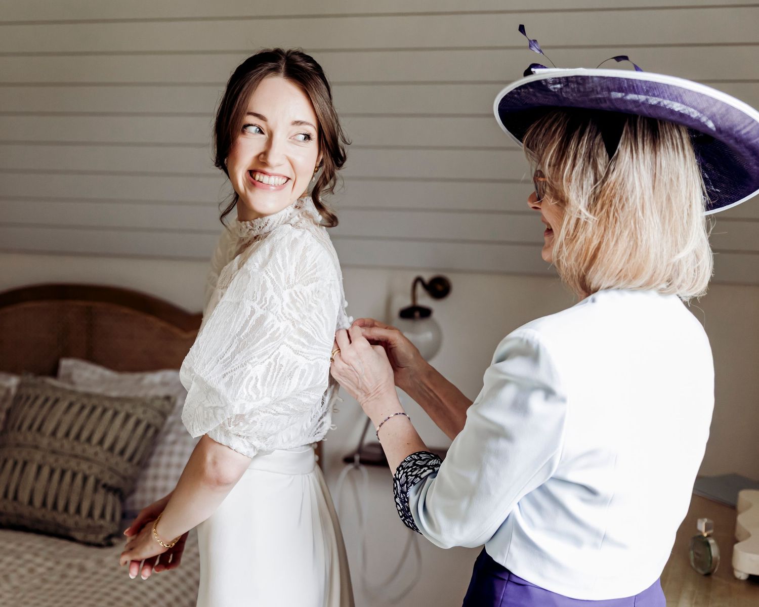 Bride smiling as woman in purple hat helps button her white wedding dress in a bright bedroom.