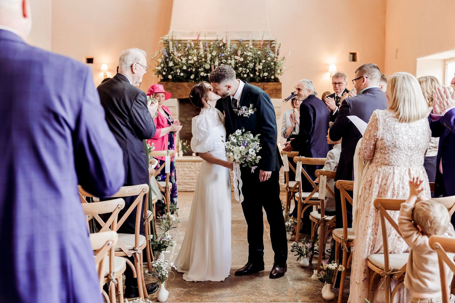 Bride and groom share first kiss at wedding ceremony aisle surrounded by guests and floral decorations.