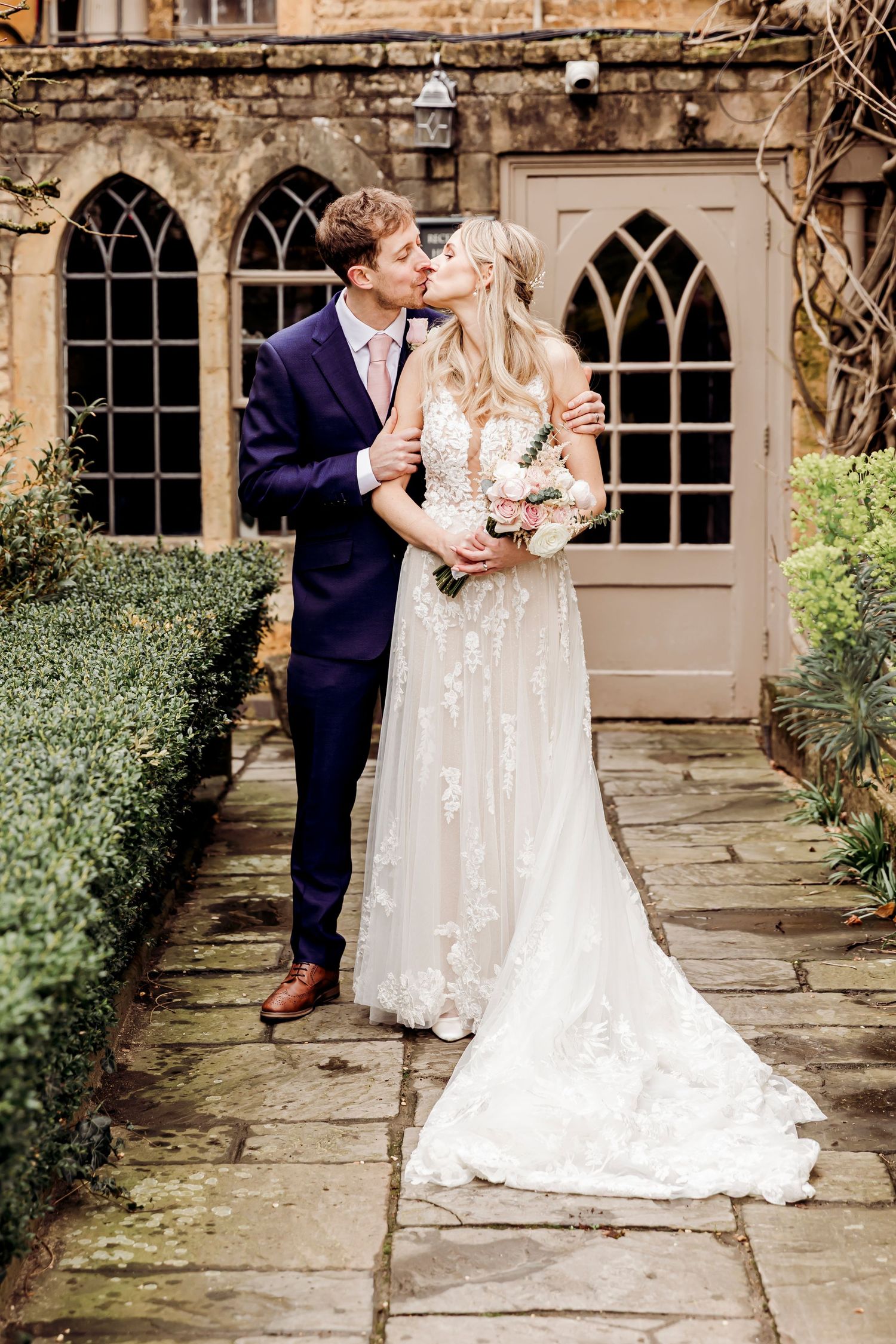 Bride and groom sharing a kiss on a stone pathway outside a historic venue with Gothic windows.