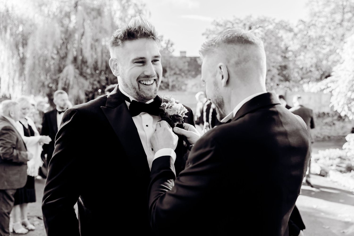 Black and white photo of groom smiling as groomsman adjusts his boutonniere at outdoor wedding.