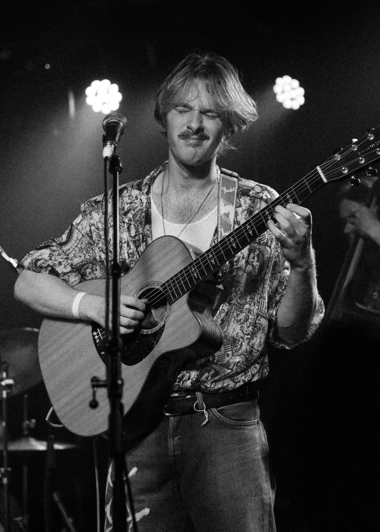 Young man with mustache smiling while playing acoustic guitar on stage in black and white concert photo.